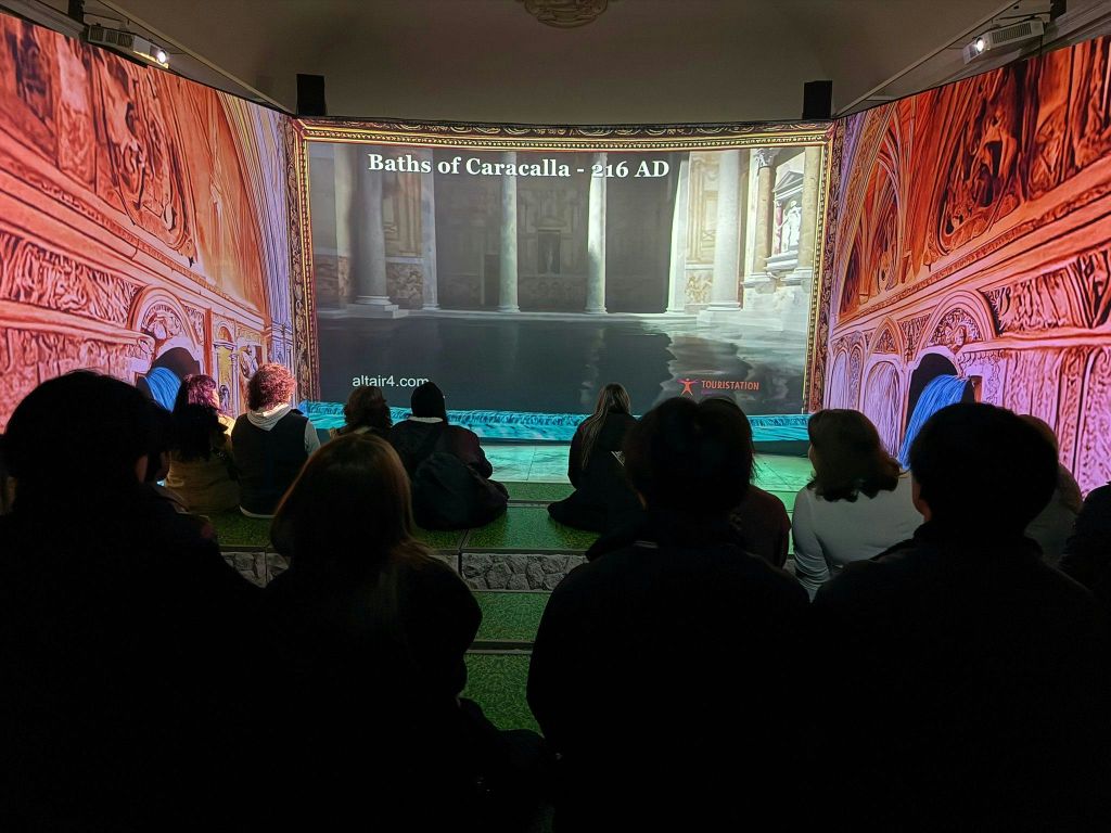 실내에 앉아 서기 216년 카라칼라 목욕탕의 투영을 지켜보는 사람들이 장식적인 주변 환경과 함께 전시되어 있습니다.