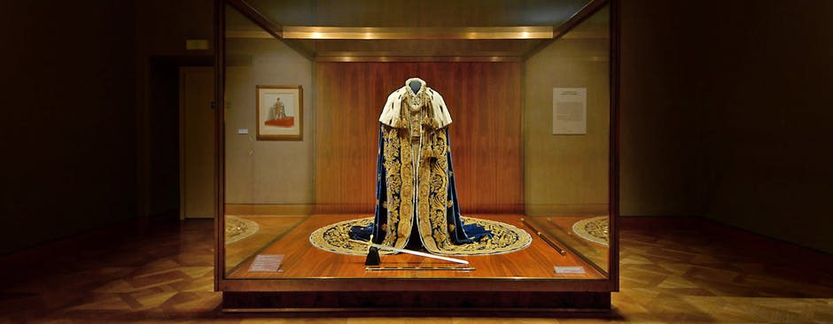 유리 케이스에 정교한 금색 자수가 진열된 왕실 가운으로, 벽에 레갈리아와 액자 문서가 함께 전시되어 있습니다.