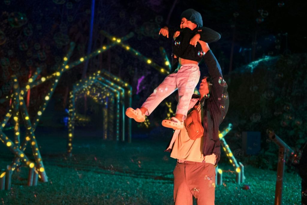 밤에 성인이 공중에서 어린이를 들어올리고, 아치형 통로에 불이 켜집니다.