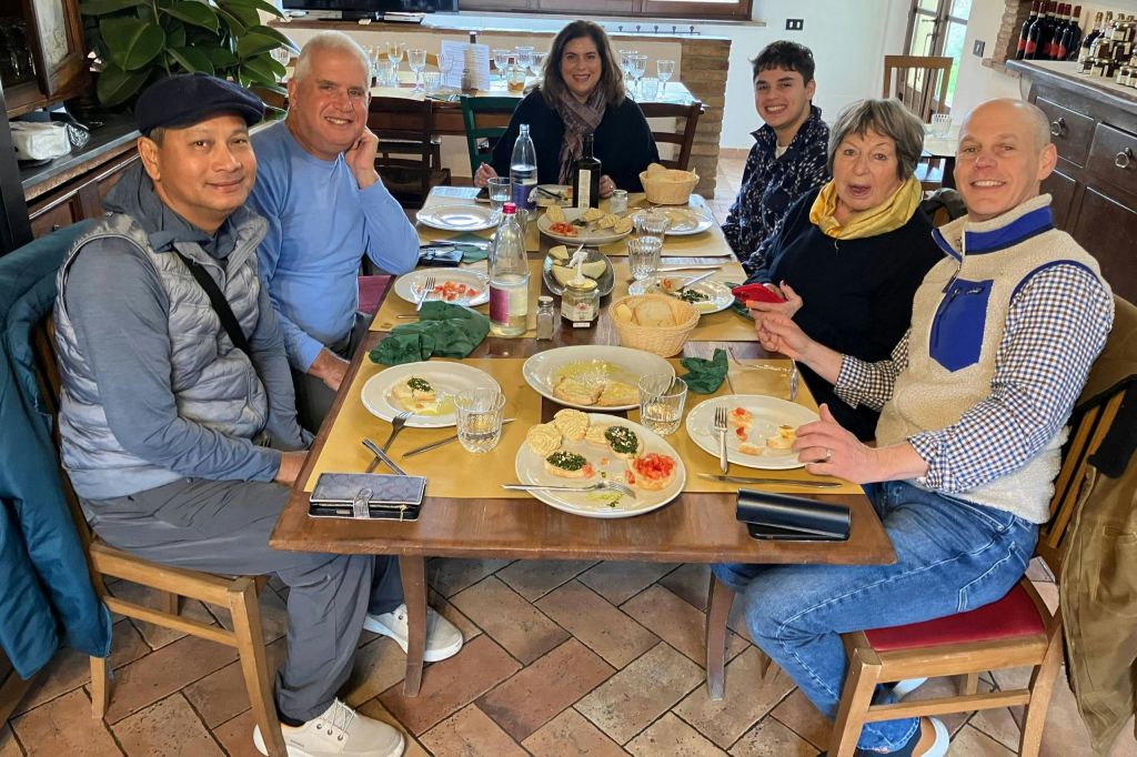 음식, 유리잔, 병이 놓인 테이블에 여섯 명이 앉아 캐주얼한 분위기에서 미소를 짓고 있습니다.