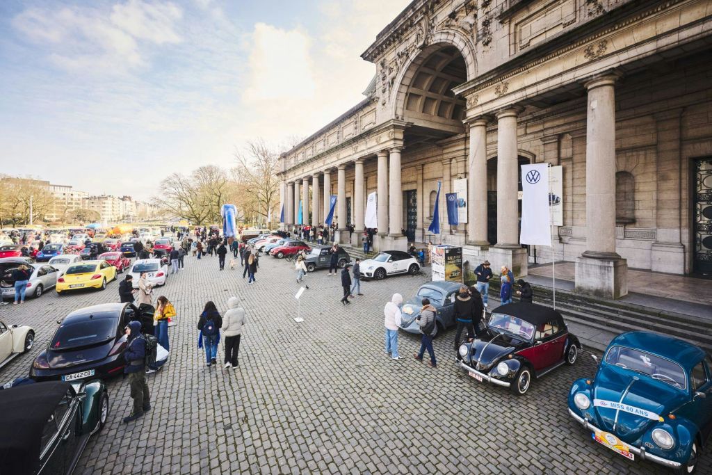 기둥과 폭스바겐 배너가 있는 크고 유서 깊은 건물 앞에서 빈티지 자동차를 둘러보는 사람들.