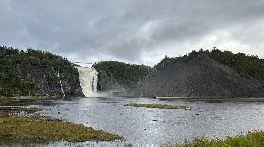 커다란 폭포가 절벽을 따라 강으로 이어지며, 위에 현수교가 보이고 머리 위로 흐린 하늘이 보입니다.