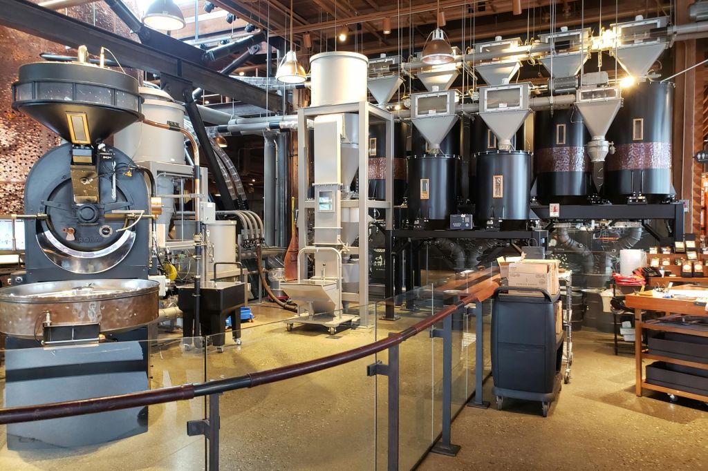 대형 금속 사일로, 파이프 및 기계를 갖춘 산업용 커피 로스팅 시설로 유리 장벽과 노출된 목재 천장이 있습니다.