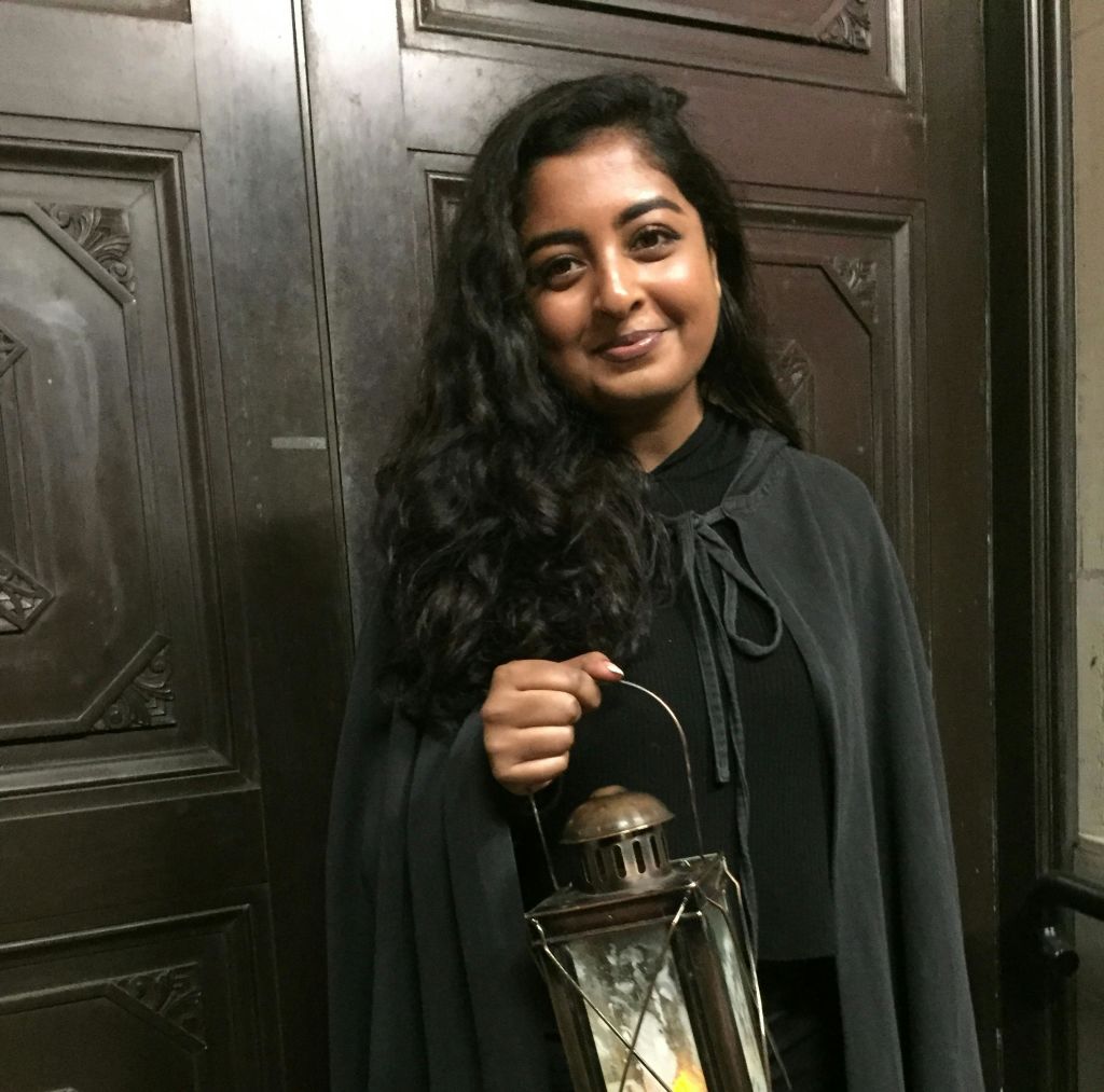 어둡고 물결 모양의 머리카락을 가진 사람이 장식 패널이 있는 커다란 나무 문 앞에 서서 랜턴을 들고 있습니다.