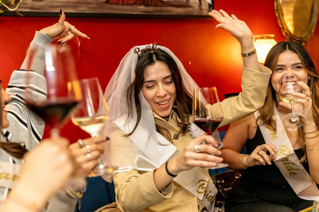 신부 베일과 띠를 두른 여성이 축제 분위기에서 와인잔을 들고 친구들과 축하합니다.