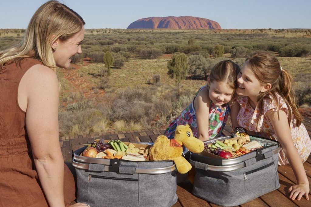 커다란 바위층을 배경으로 사막 풍경 속에서 두 어린이를 포함한 세 사람이 바구니에 음식을 가득 담고 피크닉을 즐깁니다.