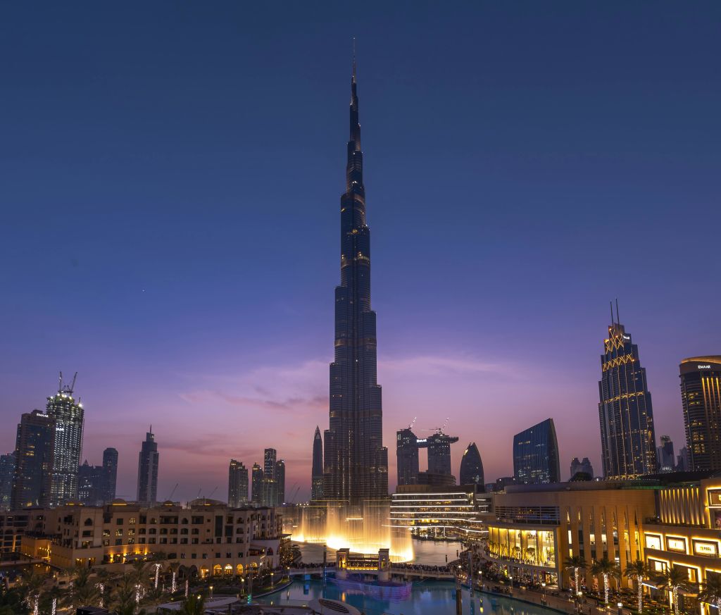 해질 무렵 도시 풍경 속에 높이 솟은 조명이 켜진 고층 건물이 서 있으며, 주변 건물과 전경에 반사된 수영장이 있습니다.