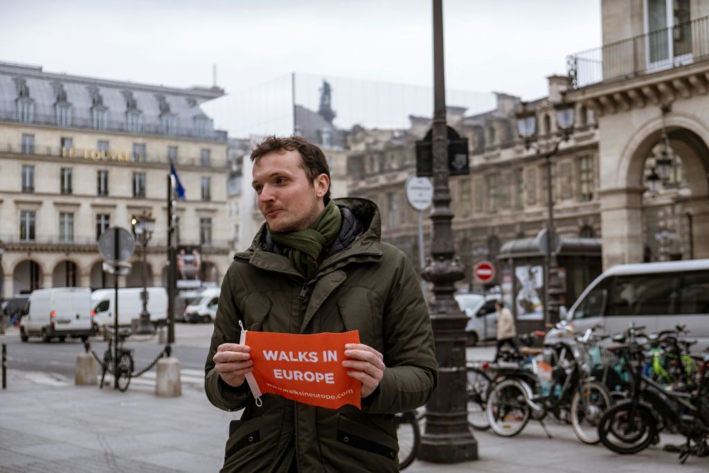 "Walks in Europe" 이라고 적힌 빨간 간판을 들고 있는 녹색 코트를 입은 사람이 건물과 자동차가 있는 유럽 거리에 서 있습니다.