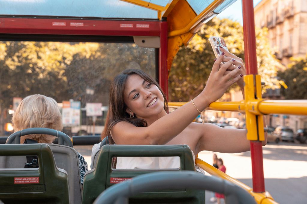 나무가 배경으로 보이는 오픈탑 버스에서 셀카를 찍으며 웃고 있는 젊은 여성.