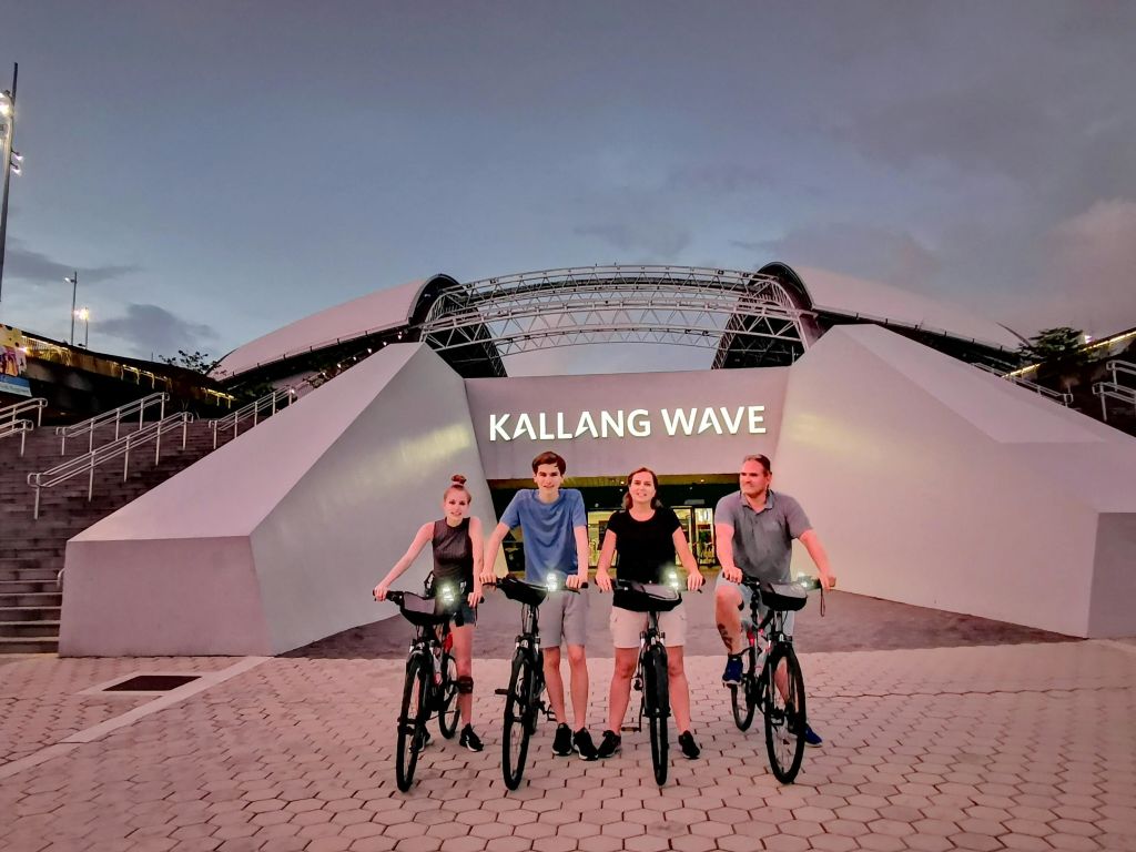 해질 무렵 칼랑 웨이브 쇼핑몰 입구 앞에서 자전거를 탄 네 명이 포즈를 취하고 있습니다.