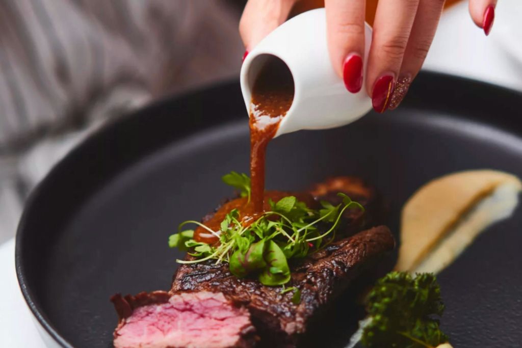 빨간 매니큐어를 바른 손이 흰색 주전자의 소스를 검은색 접시에 그린으로 장식된 스테이크에 붓습니다.