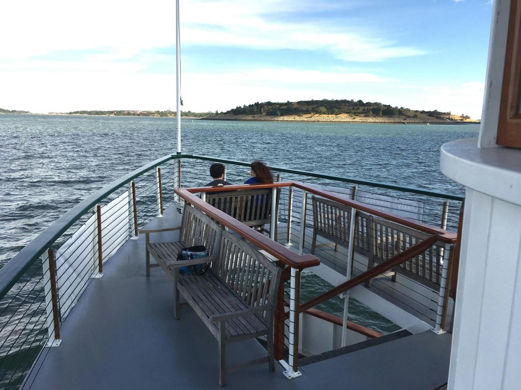 보트 갑판의 벤치에 앉아 있는 두 사람은 부분적으로 흐린 하늘 아래 먼 섬과 잔잔한 물을 마시고 있습니다.