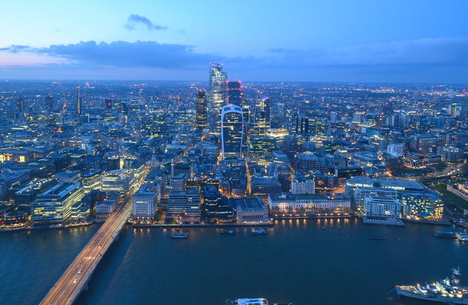 조명이 켜진 건물, 강, 왼쪽에 다리가 있는 해질 무렵의 도시 풍경을 공중에서 감상할 수 있습니다.