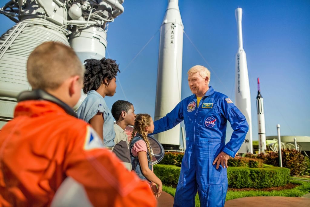 화창한 날 로켓을 배경으로 아이들과 대화하는 푸른 NASA 우주복을 입은 남성.