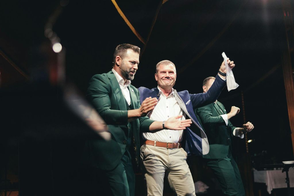 양복을 입은 세 남자가 희미하게 빛나는 무대에서 춤을 추며 박수를 치고, 한 남자는 공중에 천을 들고 있습니다. 이 장면은 축하할 만한 장면으로 보입니다.