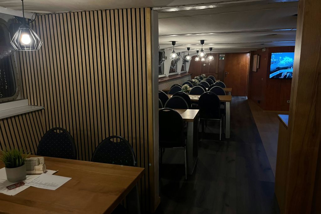 나무 테이블과 의자가 줄지어 있고, 장식용 화분과 매달려 있는 조명기구가 있는 어두운 내부입니다.