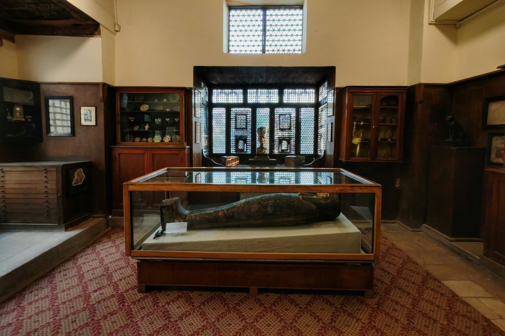 유물 선반과 창문을 배경으로 유리 케이스에 고대 목조 관을 전시한 박물관 방.