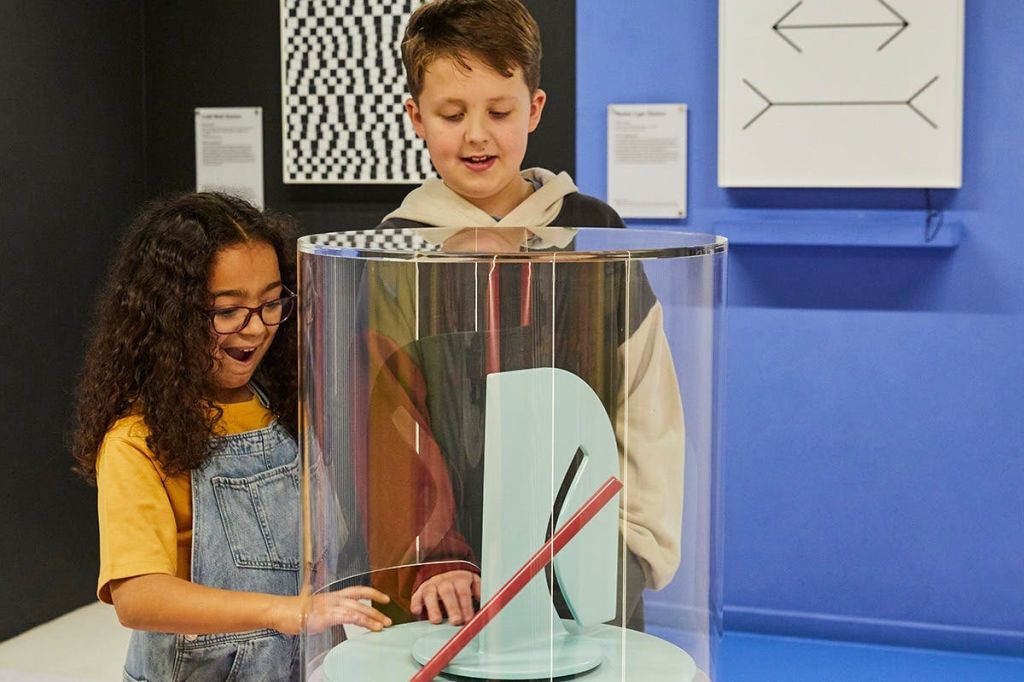 두 어린이가 박물관에서 유리로 둘러싸인 전시물과 상호 작용합니다. 한 아이는 놀란 표정을 짓고, 다른 아이는 미소를 짓습니다.