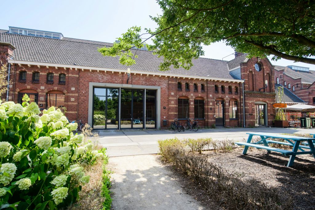 커다란 창문이 있는 붉은 벽돌 건물, 외부에 주차된 자전거, 꽃과 나무가 있는 정원으로 이어지는 통로.