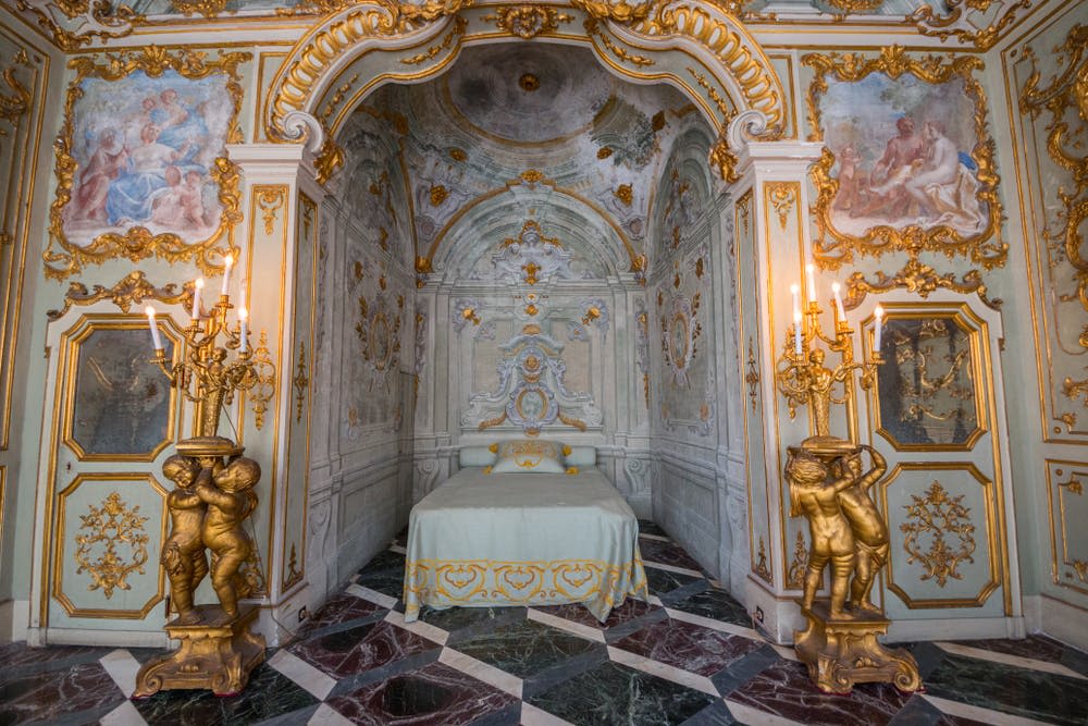화려한 골드 디테일, 캐노피 침대, 대리석 바닥, 그룹 조각상이 보유한 황금색 칸델라브라가 있는 호화로운 로코코 스타일 객실입니다.