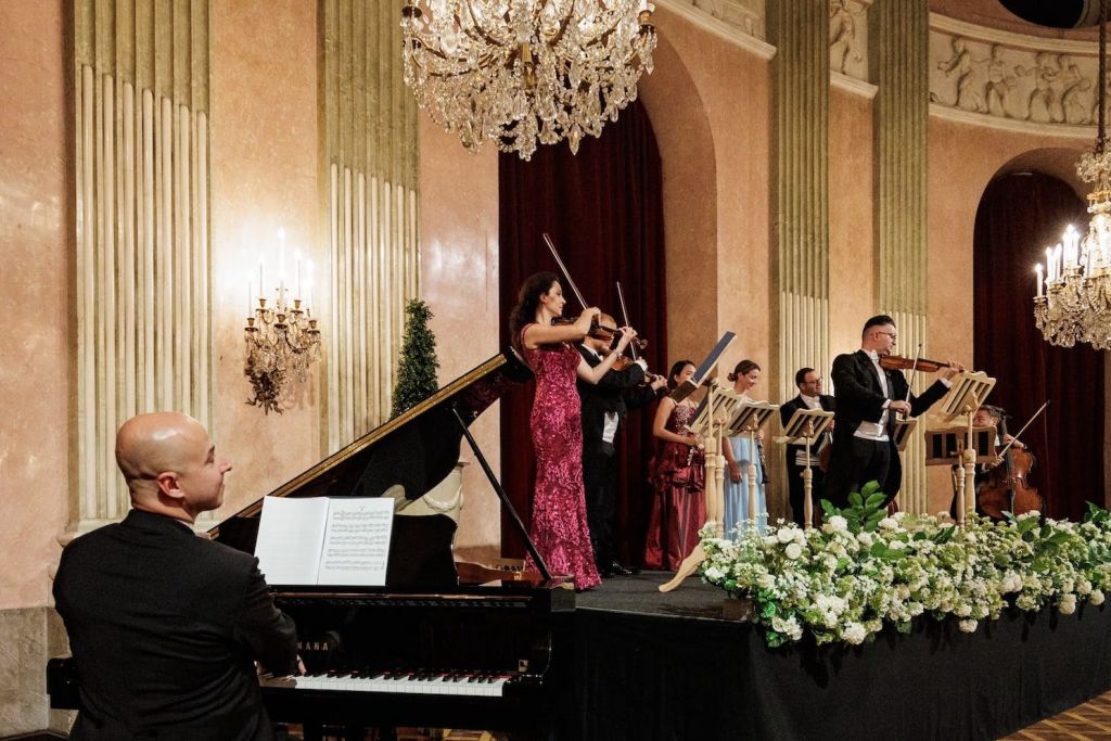 클래식 음악 앙상블이 무대에서 현악기로 연주되며, 샹들리에와 꽃꽂이로 장식된 화려한 홀에서 공연됩니다.