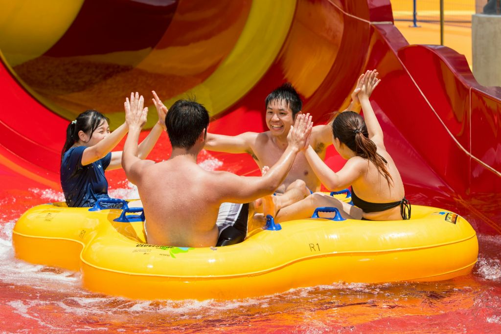 워터파크에서 노란색 팽창식 뗏목을 타고 수영복을 입은 세 사람이 화려한 워터 슬라이드 근처에서 하이파이브를 하며 미소를 짓고 있습니다.