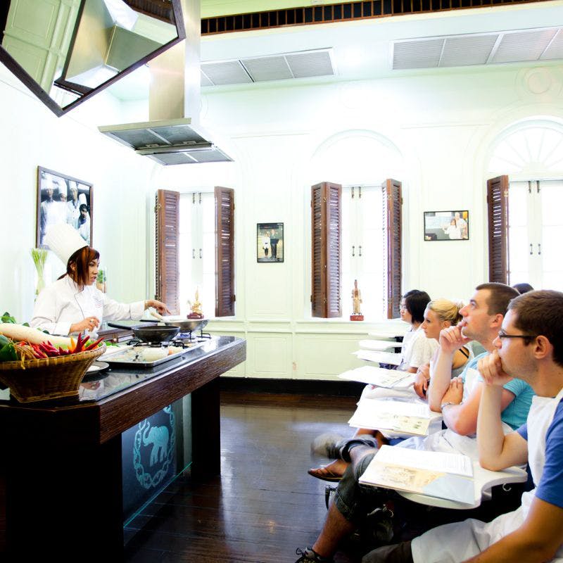 조명이 밝은 주방 교실에서 자리에 앉은 세심한 학생들에게 요리 기법을 시연하는 셰프.