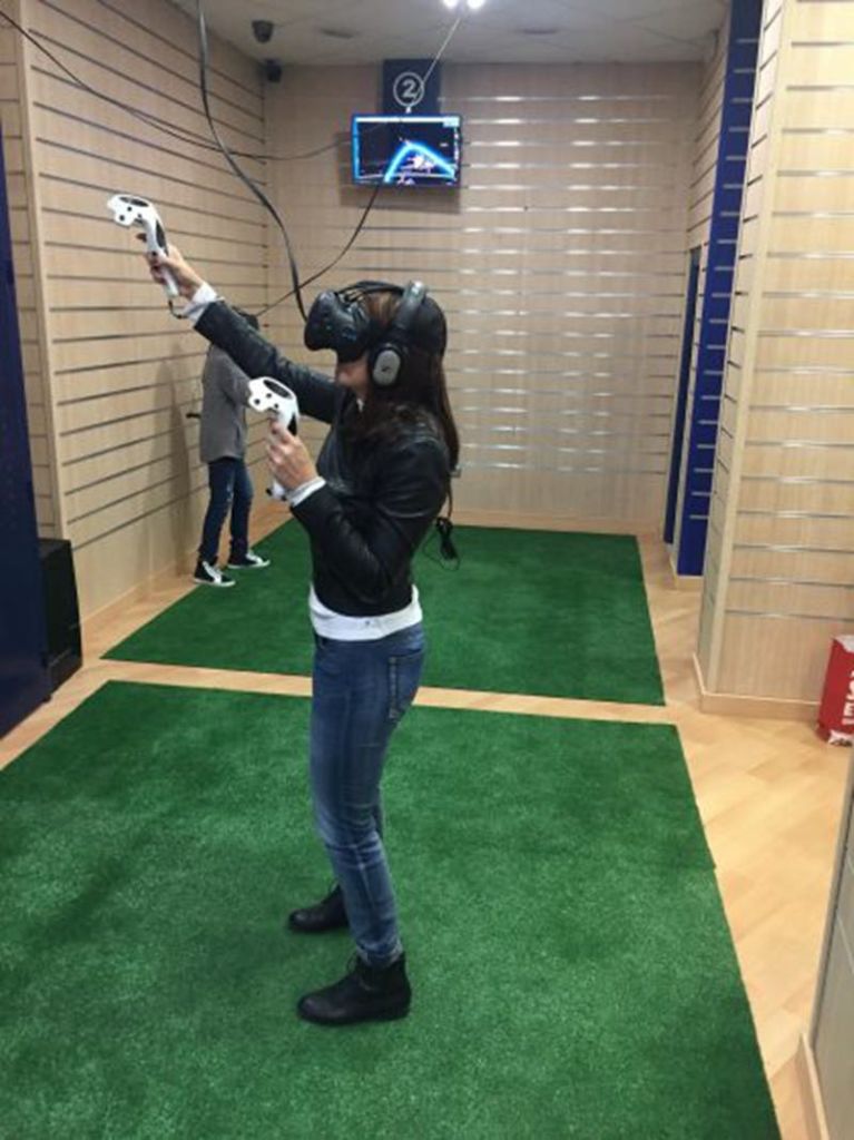 VR 헤드셋을 착용하고 컨트롤러를 들고 방의 녹색 카펫 위에 서 있는 사람. 다른 사람이 배경에 있습니다.