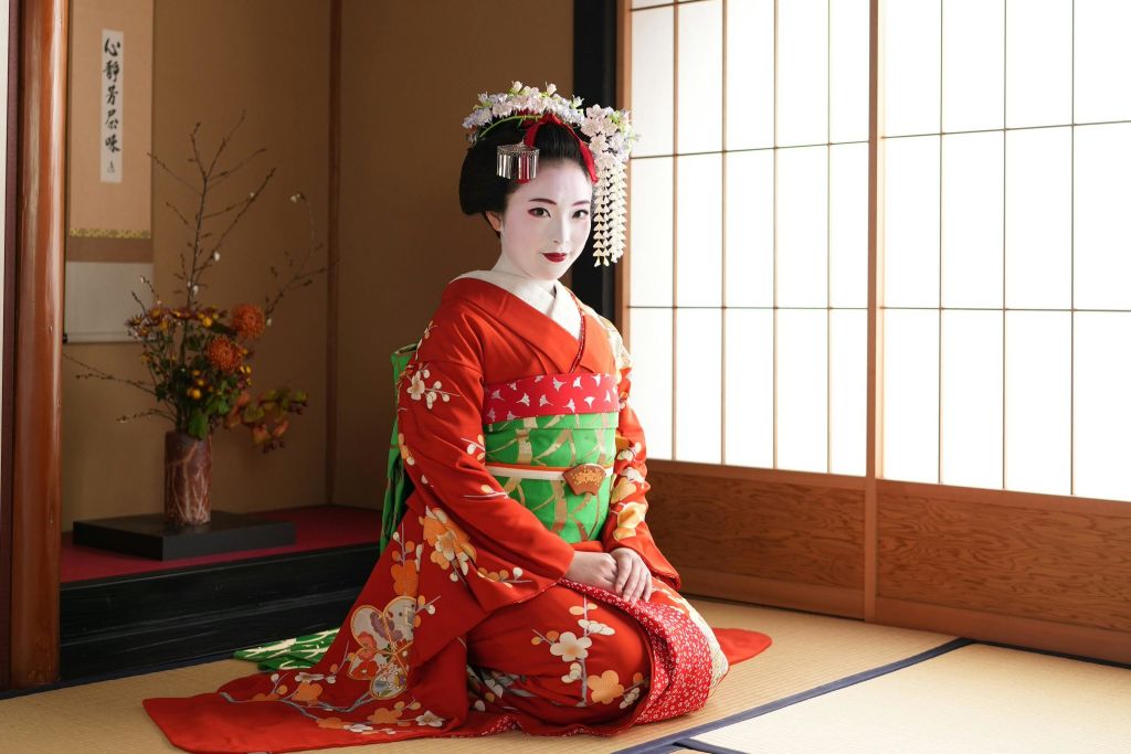 빨간색 기모노와 꽃무늬 헤드피스를 포함한 일본 전통 복장을 한 여성이 쇼지 문이 있는 방의 다다미 매트에 무릎을 꿇고 있습니다.