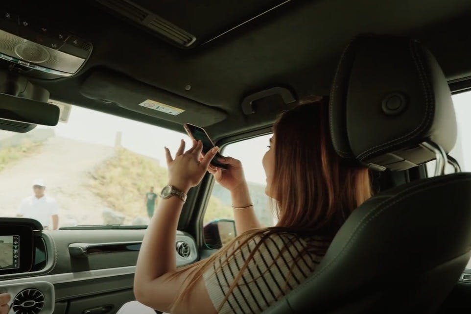 자동차에 앉아 있는 사람이 앞 유리창을 통해 사진을 찍고 있습니다. 산 중턱 근처에서 흰 옷을 입은 다른 사람이 밖에서 보입니다.