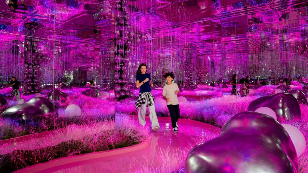 선명한 분홍색 조명, 거울 벽, 장식적인 반사 요소가 있는 방을 걷는 두 사람.