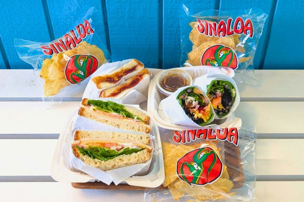 테이블 위에 브랜드가 새겨진 "시날로아" 봉지에 모듬 샌드위치, 랩, 칩. 파란색 배경.