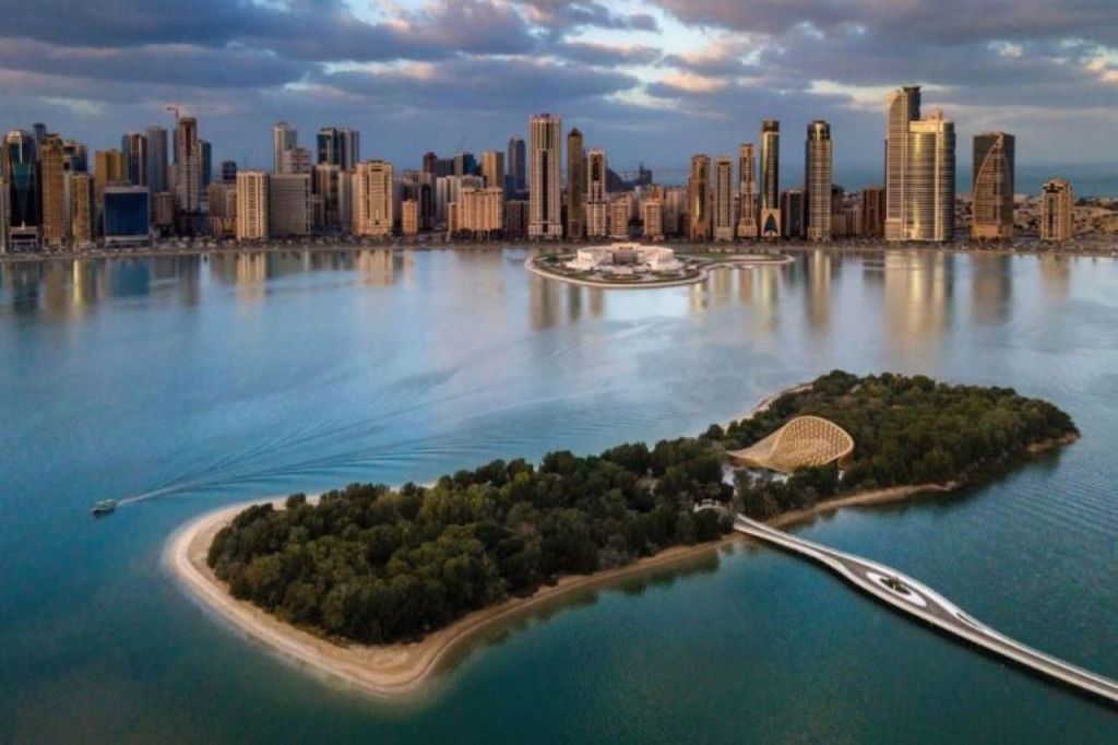 울창한 녹음과 나무 파빌리온이 있는 섬으로, 부분적으로 흐린 하늘 아래 물줄기를 가로지르는 도시의 스카이라인과 다리로 연결되어 있습니다.