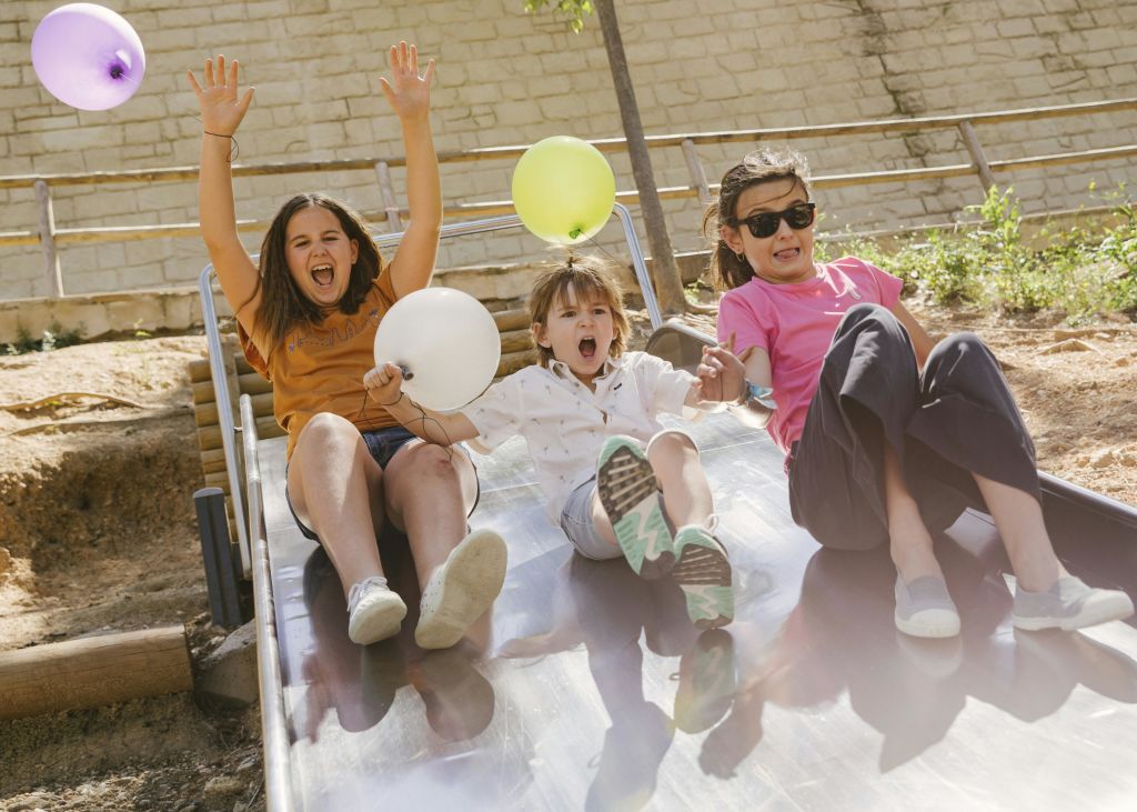 풍선을 든 세 명의 어린이가 놀이터에서 미끄럼틀을 타고 내려오며 웃으며 흥분한 표정을 짓고 있습니다.