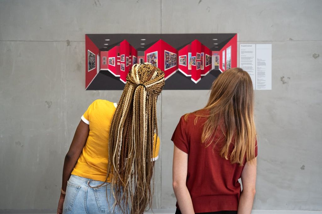 긴 머리를 한 두 여성이 빨간색과 검은색 기하학적 패턴과 사진이 돋보이는 갤러리 벽면의 액자 예술품을 관찰하고 있습니다.