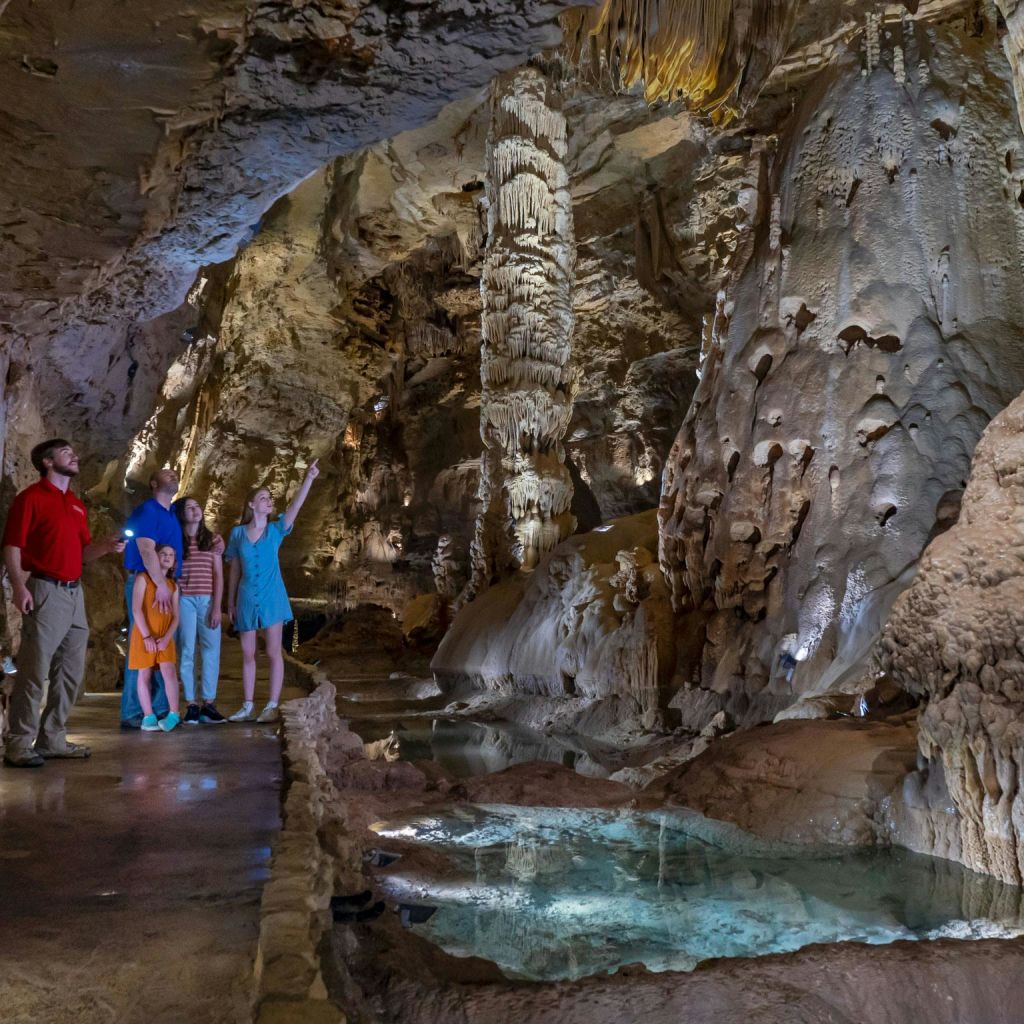 5명의 그룹이 작은 물웅덩이가 있는 조명이 밝은 동굴에서 종유석과 종유석을 관찰합니다.
