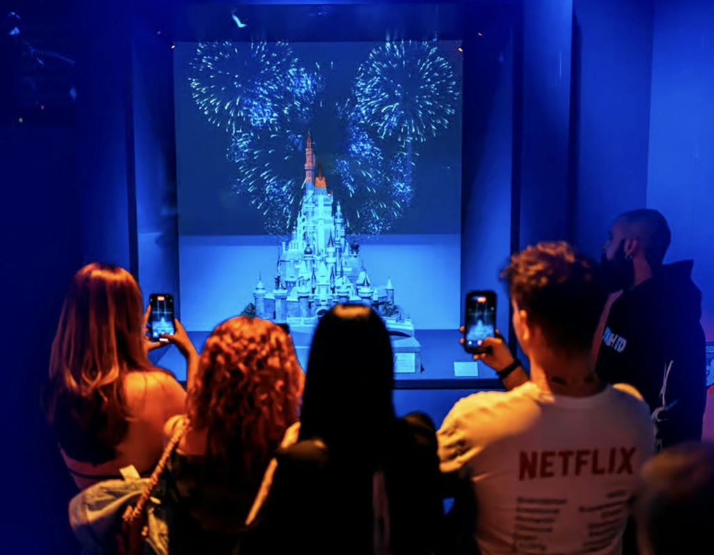 한 무리의 사람들이 조명이 희미한 배경에 불꽃이 투영된 성 모형의 전시를 보고 있습니다.