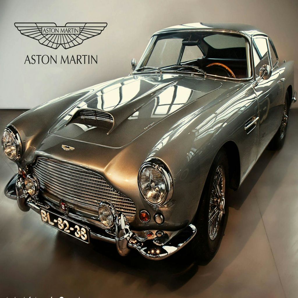 클래식 실버 Aston Martin 차량은 Aston Martin 로고와 이름을 배경으로 실내에 전시되어 있습니다.