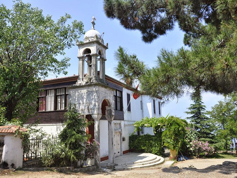 청명한 하늘 아래 나무와 녹지로 둘러싸인 아치형 입구, 종탑, 계단이 있는 작은 흰색 건물입니다.