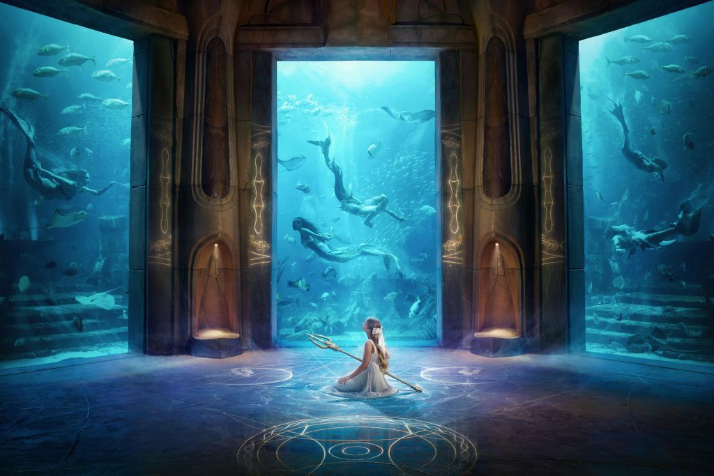 삼지창을 들고 있는 흰색 드레스를 입은 여성이 수중 테마의 방에서 빛나는 원형 패턴에 앉아 있습니다. 커다란 창문이 해양 생물을 보여줍니다.