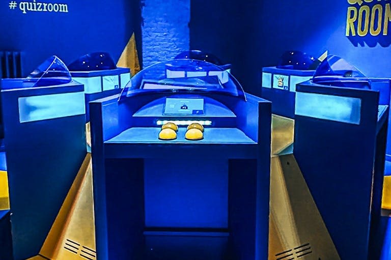 파란색 조명 방의 대화형 디스플레이로 둘러싸인 투명한 돔 아래에 노란색 신발이 전시된 박물관 전시관입니다.