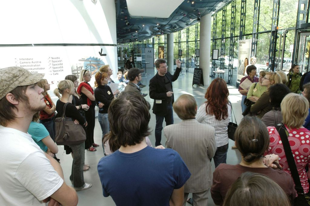 한 남성이 자연 채광이 들어오는 현대적이고 유리벽으로 마감된 실내 공간에서 한 무리의 사람들과 이야기를 나누고 있습니다.