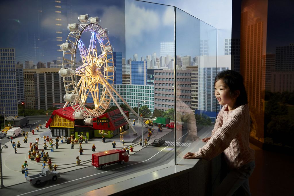 건물, 관람차, 초소형 차량이 유리 케이스에 둘러싸인 미니어처 도시 모델을 보고 있는 어린이.