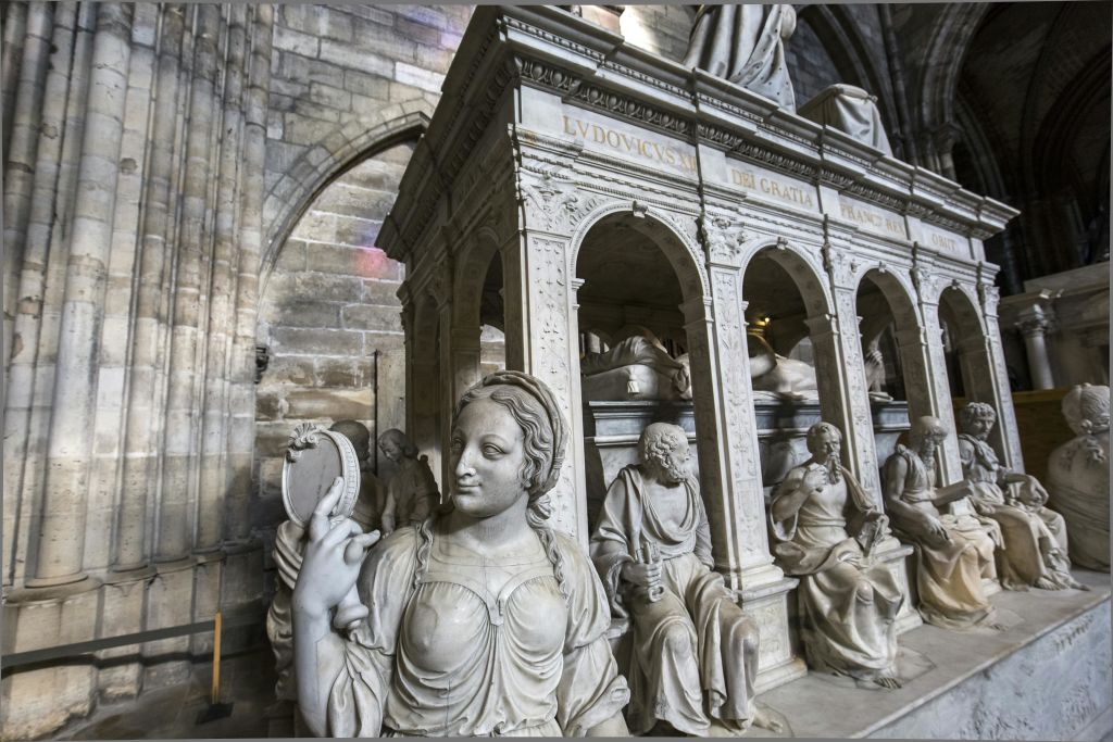 거울을 든 여성을 포함한 우아한 대리석 조각상이 석조 벽으로 둘러싸인 건물 내부의 화려한 아치형 구조물 아래에 서 있습니다.