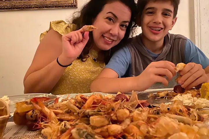 해산물 껍질로 덮인 테이블에서 한 여성과 한 소년이 새우를 껍질을 벗기고 먹으며 미소를 짓고 있습니다.