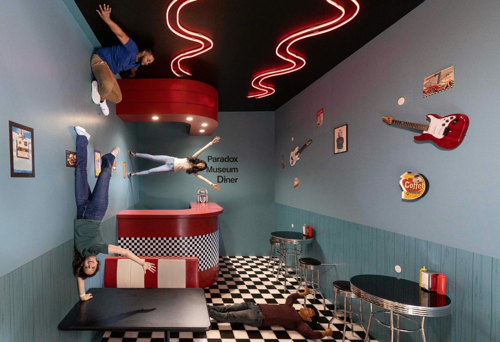 체크무늬 바닥, 빨간색 카운터, 기타로 장식된 청록색 벽이 있는 식당에서 사람들이 옆으로 서 있고 거꾸로 서 있는 것처럼 보입니다.