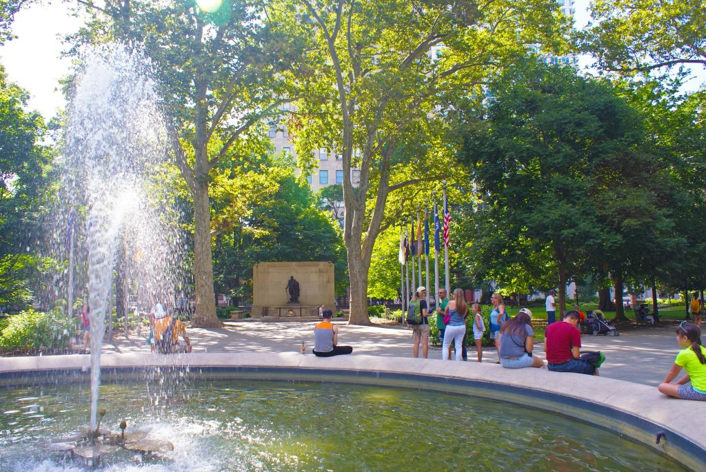 나무, 깃발, 조각상을 배경으로 햇살이 내리쬐는 공원의 분수대 근처에서 휴식을 취해보세요.