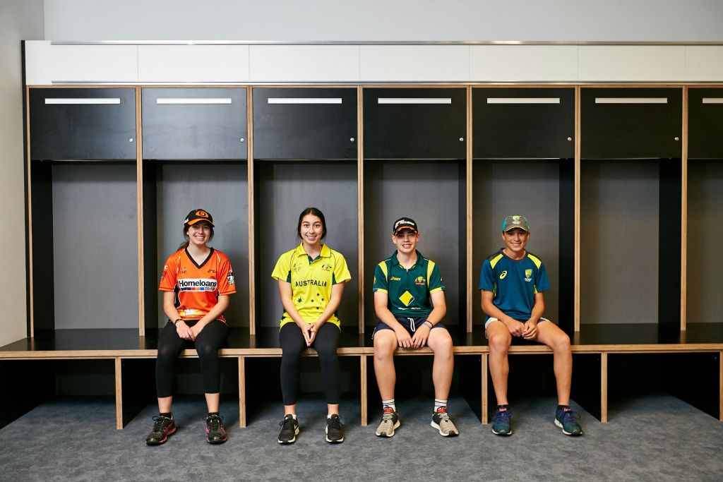 스포츠 복장을 한 네 명이 탁 트인 사물함 앞 벤치에 앉아 카메라를 보며 미소를 짓고 있습니다.