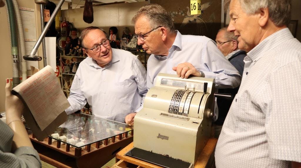 세 명의 노인이 다양한 수집품이 있는 방에서 빈티지 금전 등록기를 검사합니다.