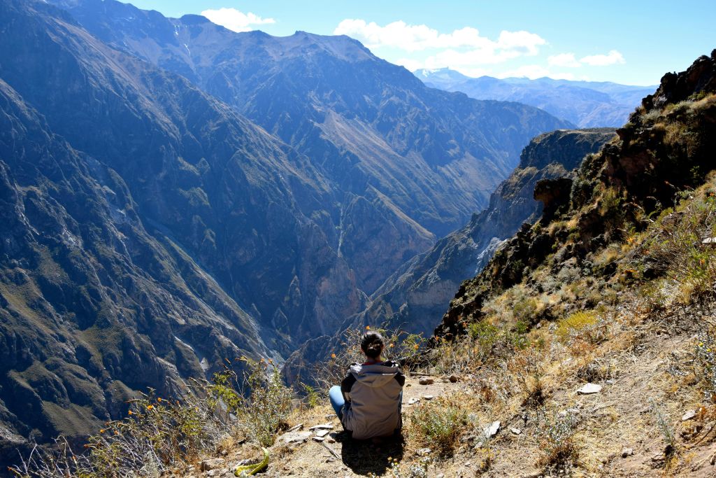 맑고 화창한 하늘 아래 광활한 산악 지형이 내려다보이는 절벽 가장자리에 앉아 있는 사람.
